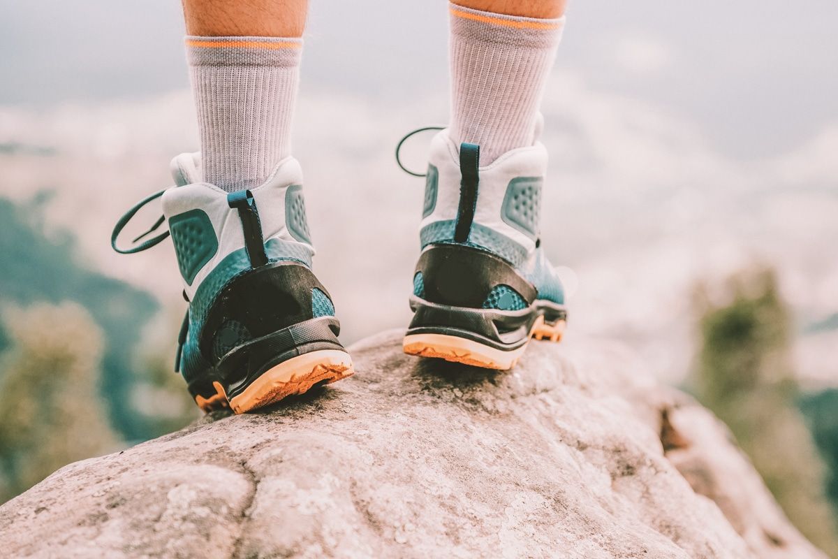 A pair of feet wearing light grey hiking socks and blue-and-white hiking boots stands on a rock overlooking an out-of-focus vista.
