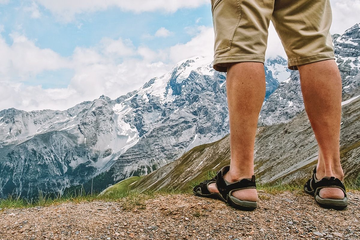 A man's feet and legs wearing black hiking sandals and khaki shorts seen from behind standing on a rocky mountaintop looking out at a snowy mountaintop vista.