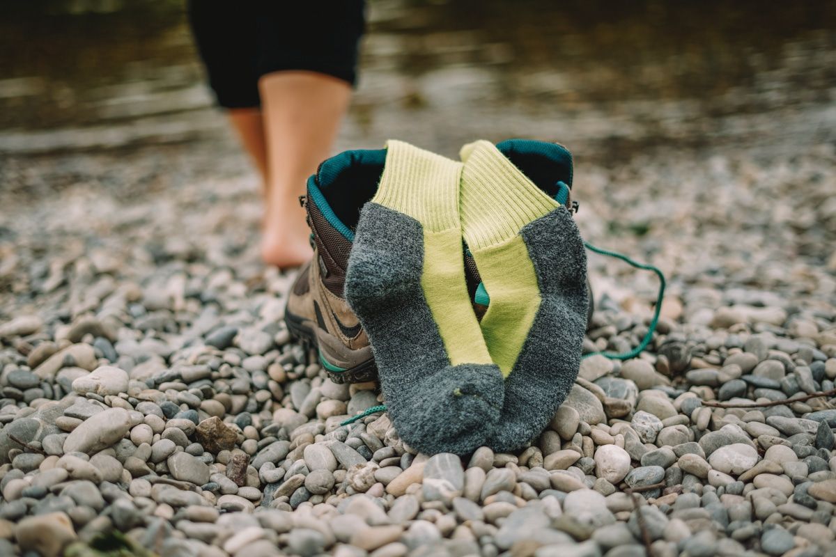 A pair of grey and neon yellow hiking socks poke out of hiking boots that are sitting on a pebbly river bank, while a pair of out-of-focus legs is visible in the background walking towards the water.