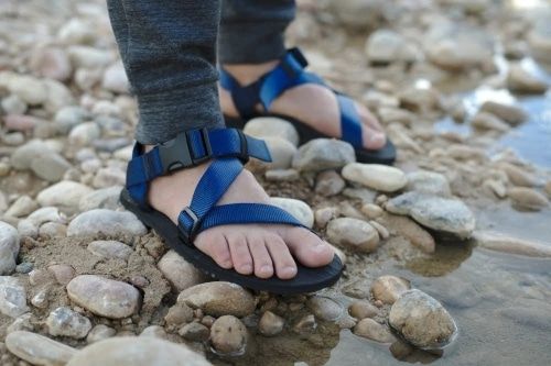 Product photo for the Unshoes Pah Tempe 2.0 in blue, modeled on a pair of feet standing on a rocky riverbank.