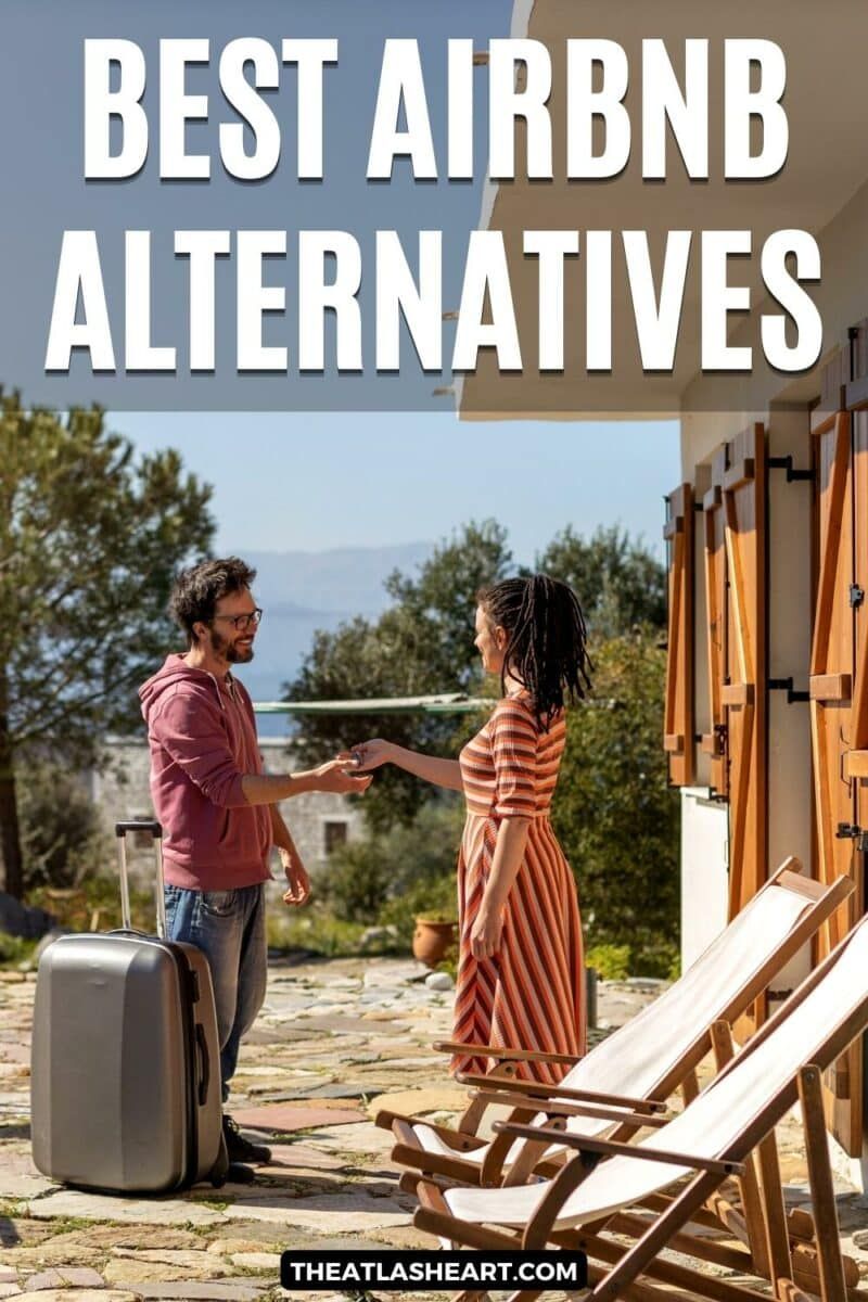 A woman in a striped dress gives a key to a man in a maroon sweatshirt and glasses with a grey hardside suitcase next to him on the stone patio outside a vacation rental, with the text overlay, "Best Airbnb Alternatives."