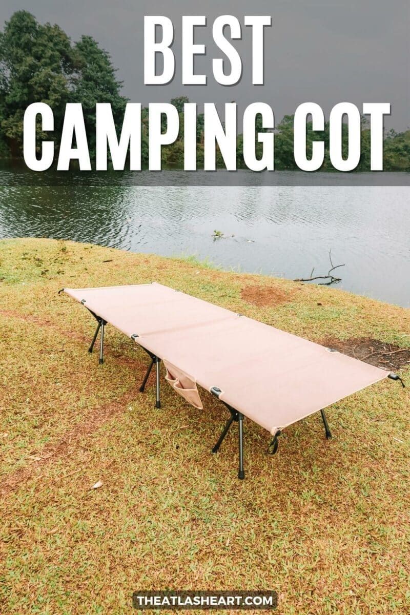 A beige canvas cot sits on a grassy bank of a greenish lake, with trees and an overcast sky in  the background, with the text overlay, "Best Camping Cot."