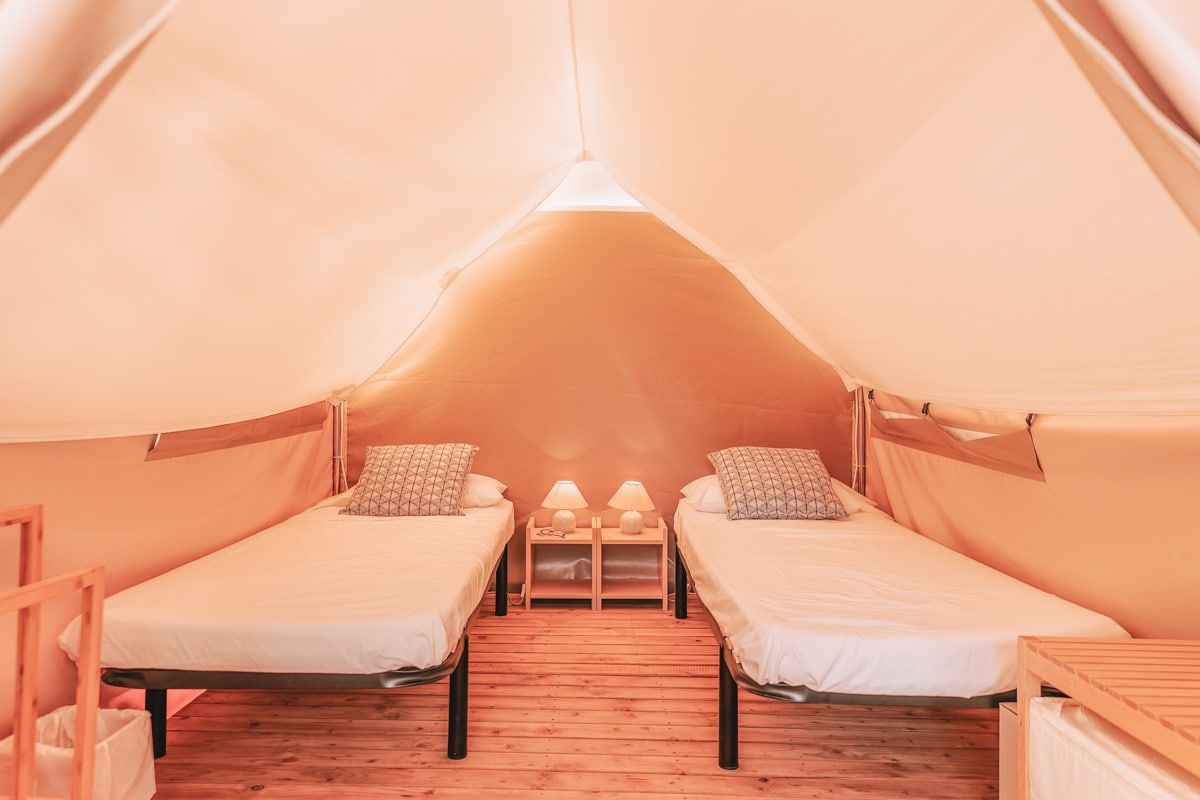 Two camping cots topped with white mattresses sitting side-by-side on a wooden platform inside a canvas glamping tent.