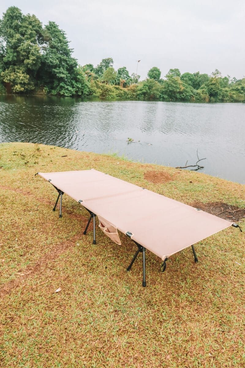 An example of one of the best camping cots, a beige canvas cot, sits on a grassy bank of a greenish lake, with trees and an overcast sky in  the background.