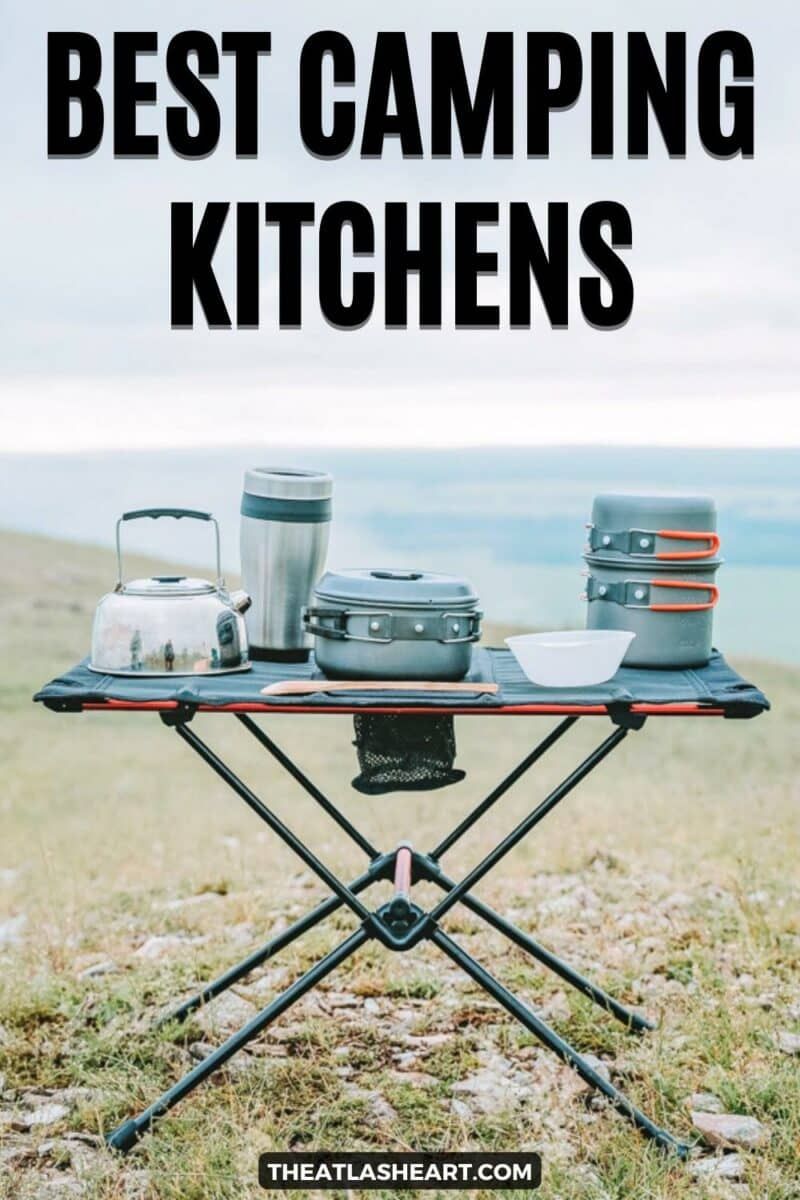 A black and orange folding camping kitchen table laid out with a mess kit sitting on a grassy hilltop with the text overlay, "Best Camping Kitchens."