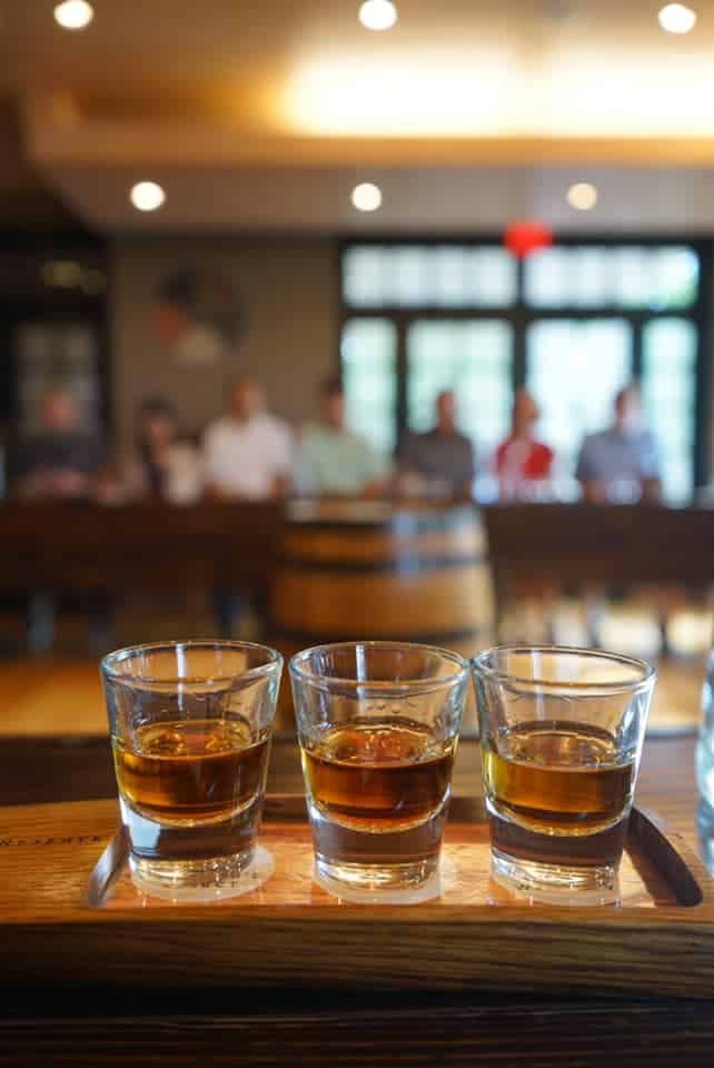 An example of one of the best gifts for bourbon lovers, three shots of bourbon lined up on a wooden surface with a soft-focus tasting room in the background.