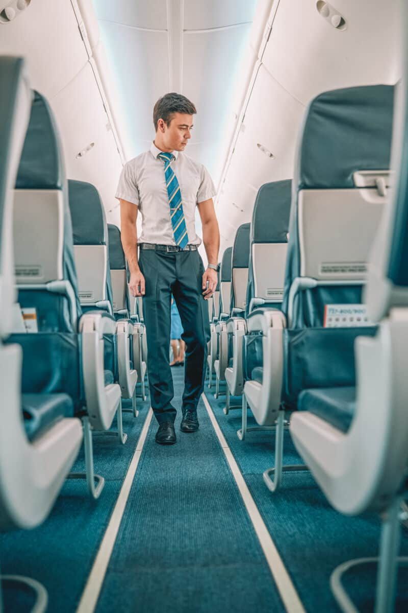 The best gifts for flight attendants: a young male flight attendant with brown hair walks down the aisle of a plane and looks at the row of seats to his left.