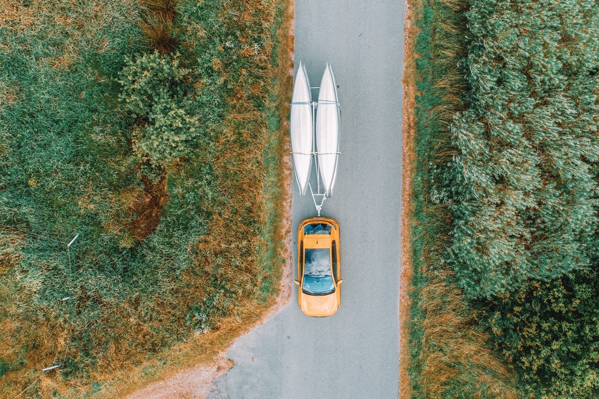 Bird's eye view of a yellow car with two white kayaks on a trailer, driving down country road lined with greenery.