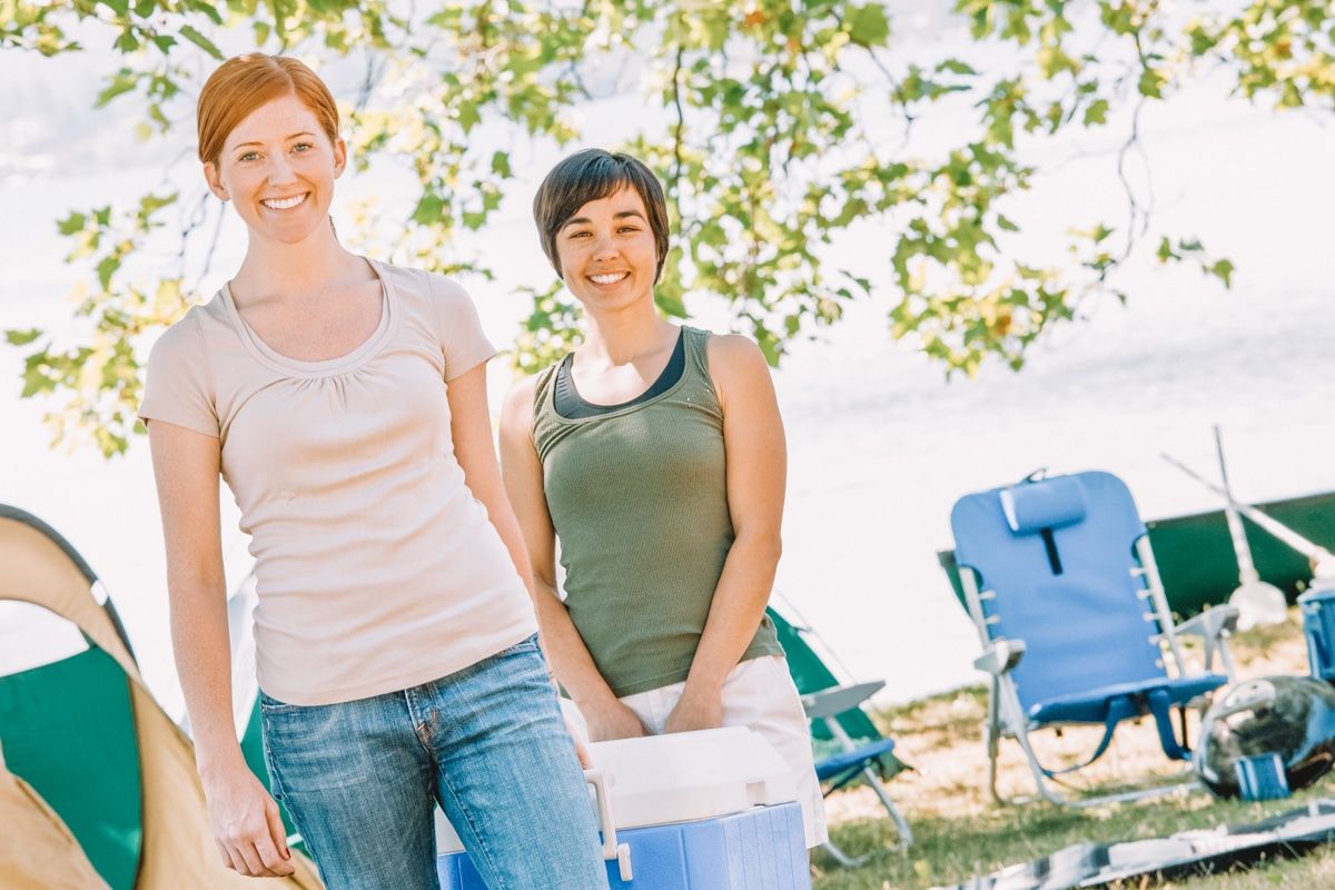 Two young women smile as they both carry a blue ice chest cooler in a sunny campsite with leafy branches behind them.