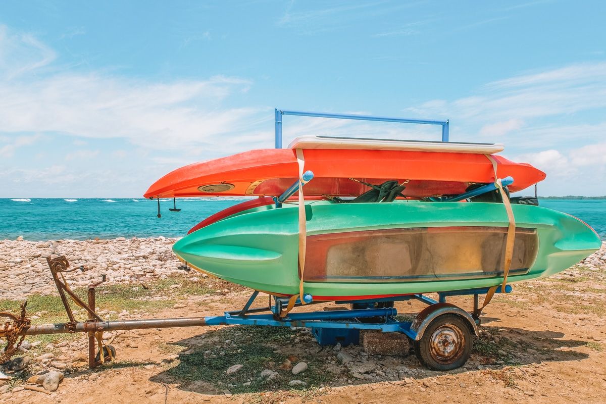 Three brightly-colored kayaks on a blue kayak trailer parked on a beach with a turquoise ocean in the background.