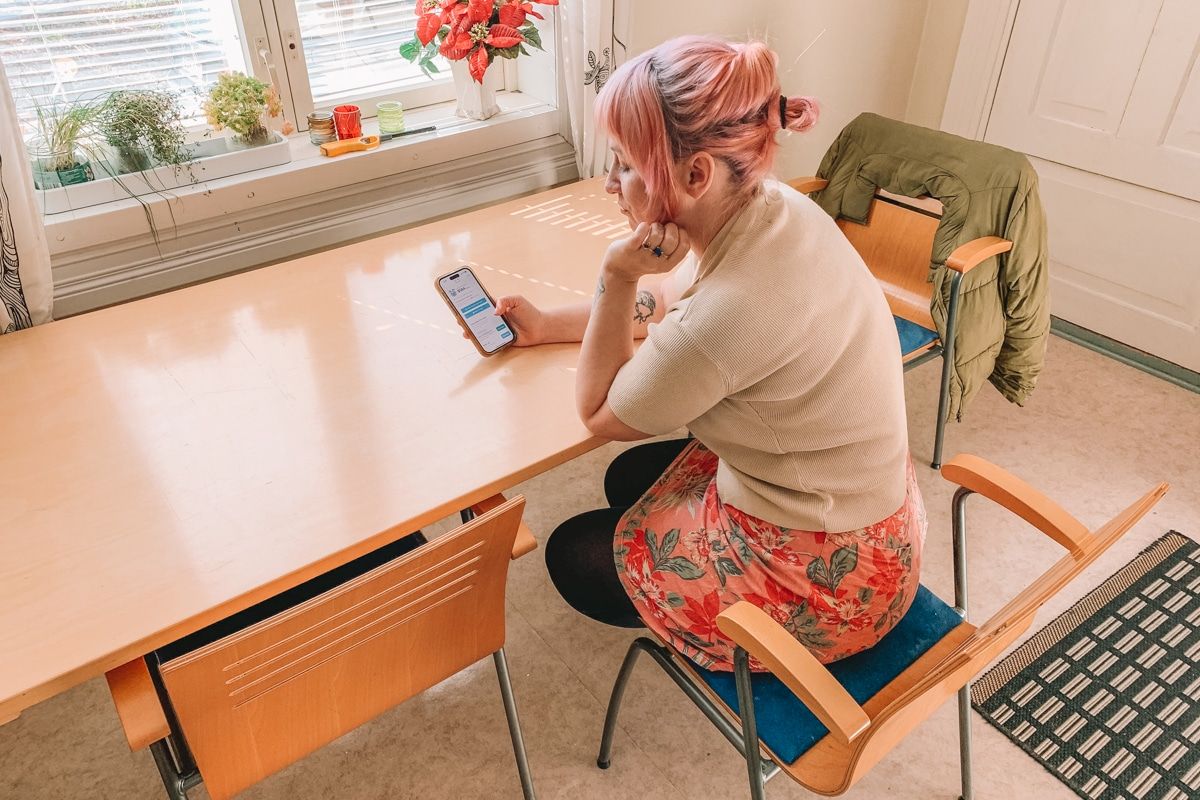 A young woman with light pink hair, wearing a pink tropical-print skirt, holds an iPhone and browses on the Hopper app while resting her elbows on a light wood kitchen table, with light coming through a window in front of her.