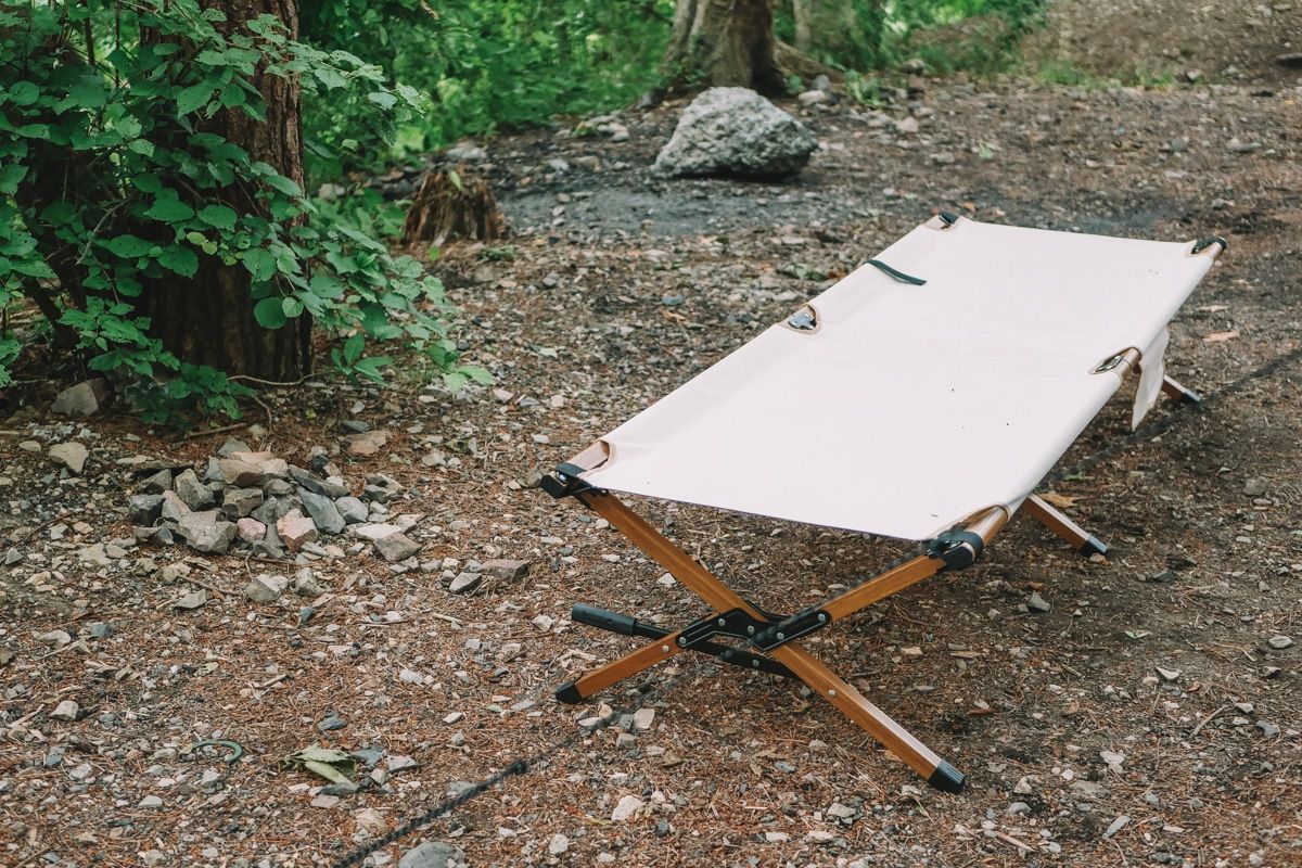 A beige canvas camping cot set up on a gravelly forest floor, with leafy vegetation surrounding.