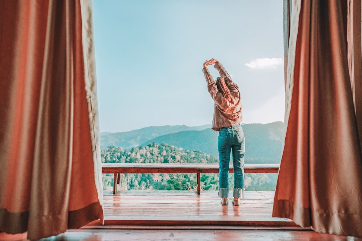 A view through parted curtains of a 
 young woman in jeans seen from behind stretching on on a wooden balcony overlooking a hilly, forested landscape.