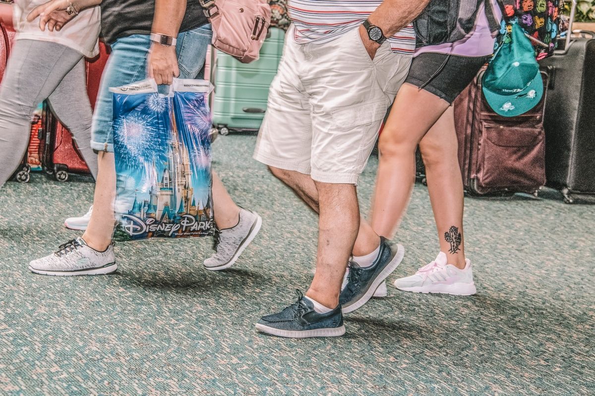 A lower view of a crowd of travelers' legs wearing walking shoes, with one person holding a Disney park shopping bag.