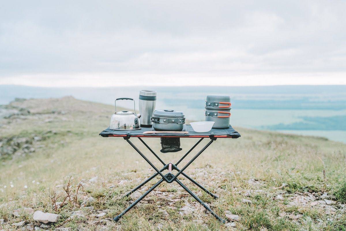 A black and orange folding camping kitchen table laid out with a mess kit sitting on a grassy hilltop with a hazy valley in the distance.