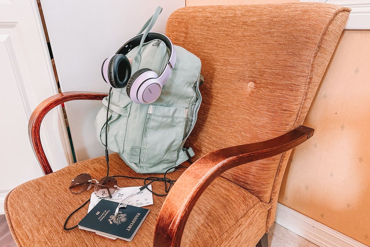 A light green backpack, lavender headphones, a pair of sunglasses, a boarding pass, and a US passport sitting on an ochre armchair with yellow wallpaper and a white armoire in the background.