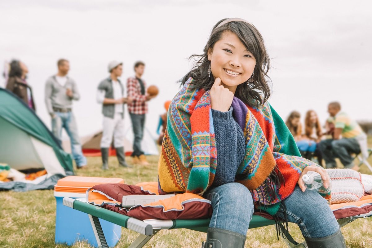A young dark-haired woman wrapped in a brightly-colored blanket smiles as she sits on a camping cot in a field, with a tent and people playing sports in the background.