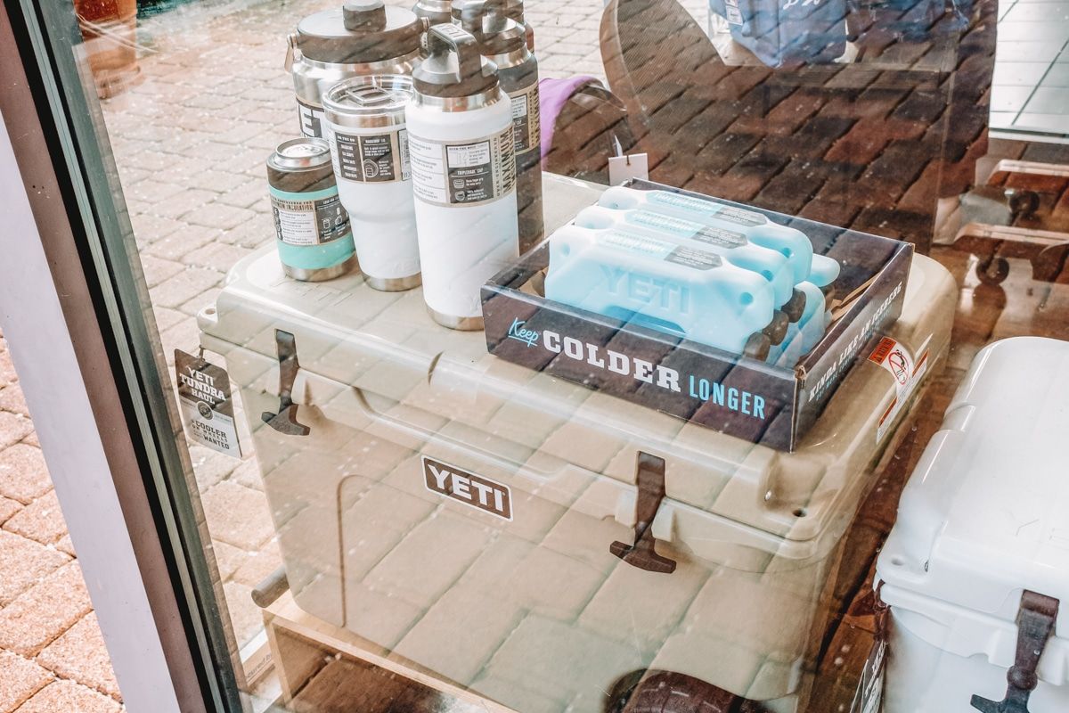A view through a glass sliding door of a tan Yeti cooler on a brick patio with several Yeti water bottles and ice packs sitting on top of it. 