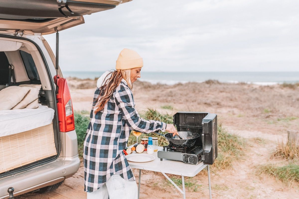 A woman with dramatic highlights and a long flannel shirt prepares a meal over a portable camping kitchen table with the open hatchback of an SUV an overcast beach scene in the background.