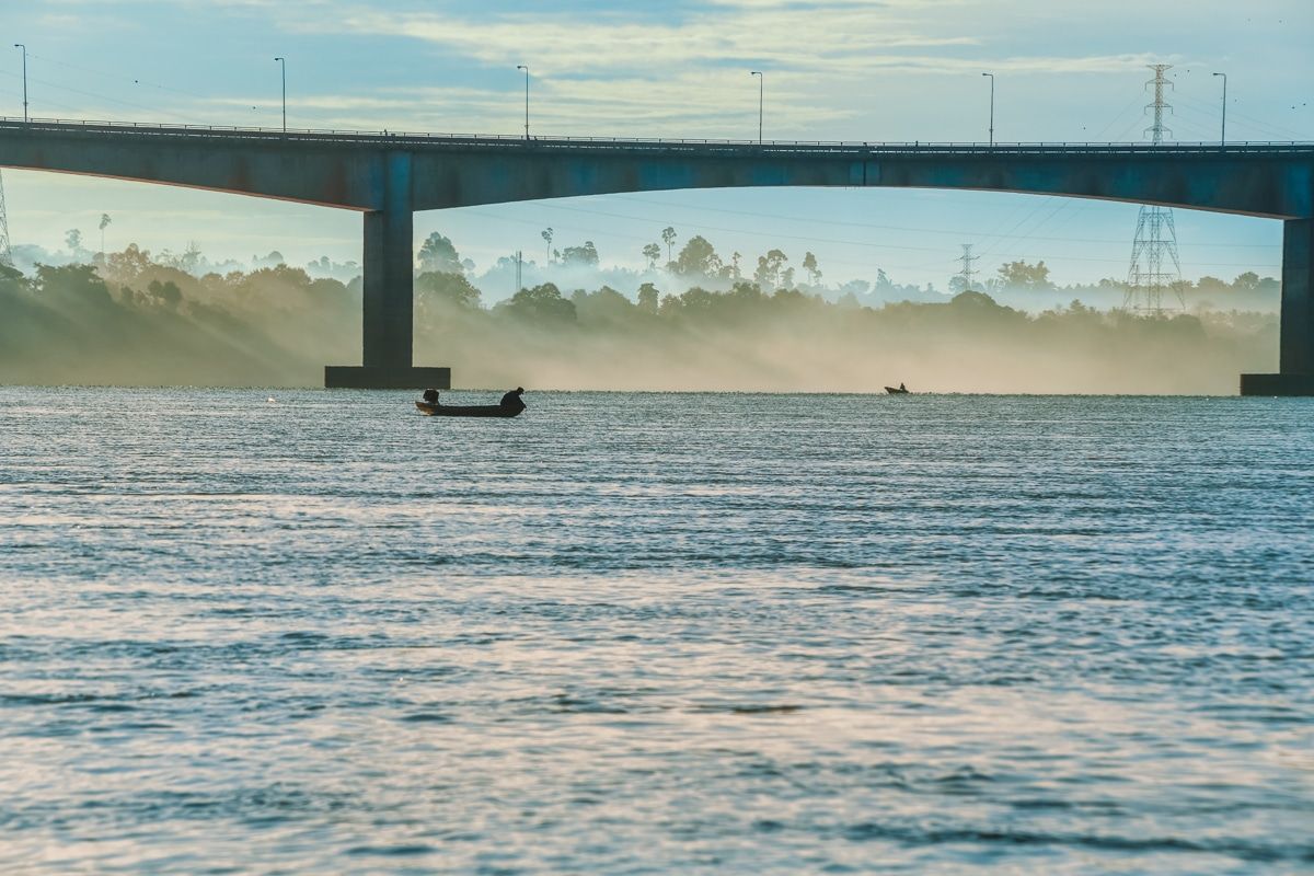 A silhouette of a fisherman in a motorized kayak on a body of water with a concrete bridge and a misty industrial landscape in the background.
