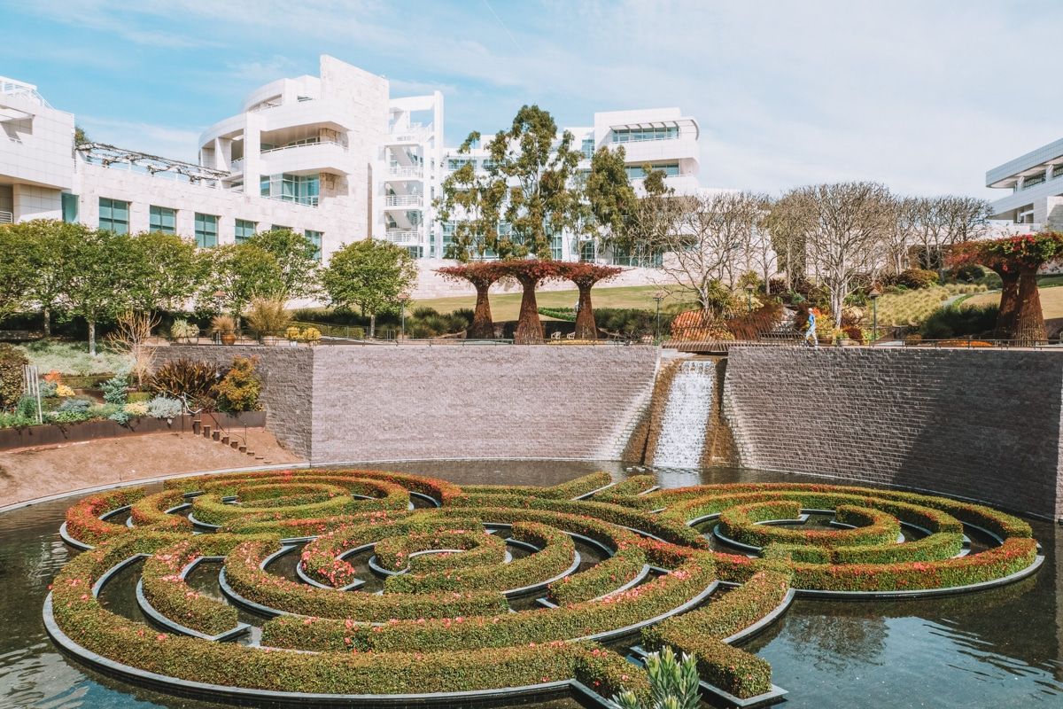 A hedge labyrinth in a pond in front of the Getty Center with a sunny, partly cloudy sky beyond.