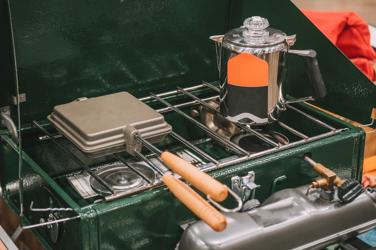 A close-up of a dark green camping stove with a coffeepot and waffle-maker sitting on it.