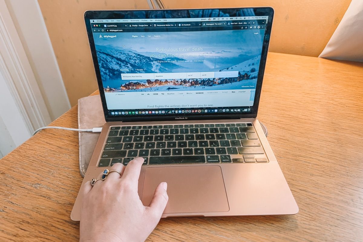 A rose gold macbook laptop displaying the Skiplagged homepage sits on a wooden desk with a yellow wall in the background with a hand scrolling on the trackpad in the foreground.