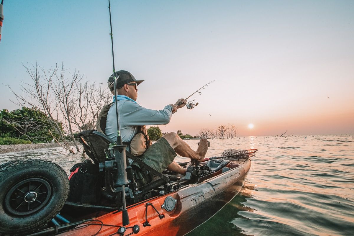 A man fishing from an orange motorized kayak on a lake with a hazy sunset in the distance.
