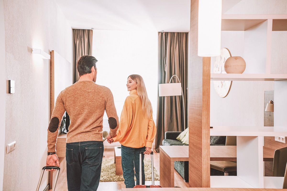 A brown-haired man and a blond woman walk hand-in-hand into a vacation rental apartment, pulling rolling suitcases, with bright light coming through the living room window behind them.