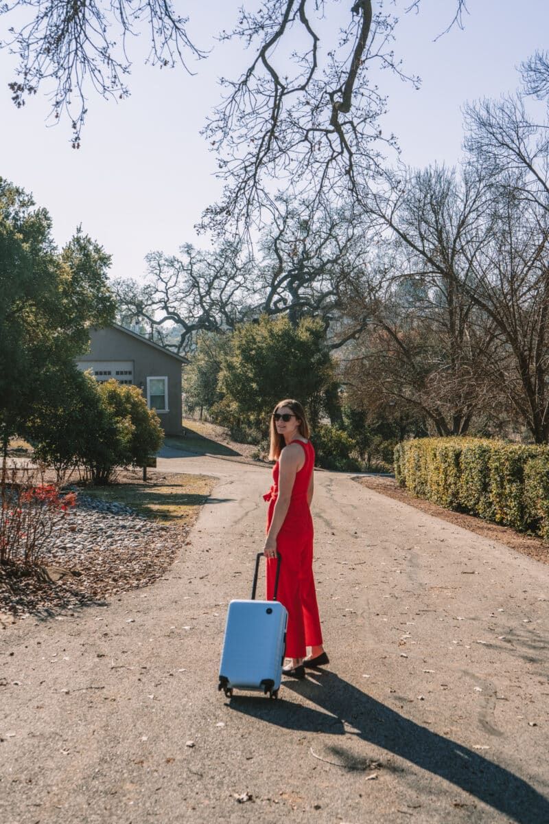 A young woman in a red jumpsuit and sunglasses looks back over her shoulder as she pulls a piece of one of the best hardside luggage sets, a light blue carry-on roller, down a long, wooded driveway in afternoon sun.