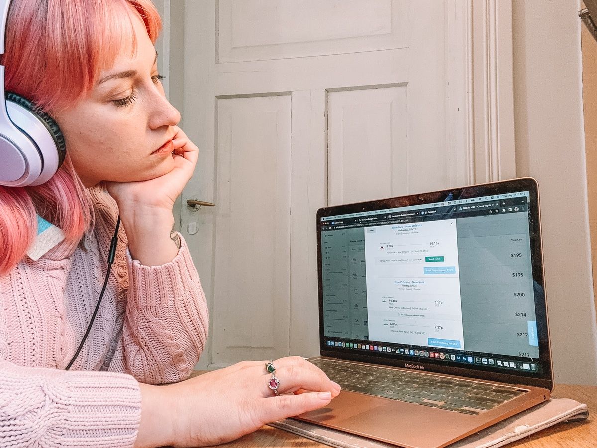 A woman with pink hair wearing lavender headphones and a pink sweater sits at a desk and looks at the Skiplagged homepage on her laptop, with a closed white door in the background.