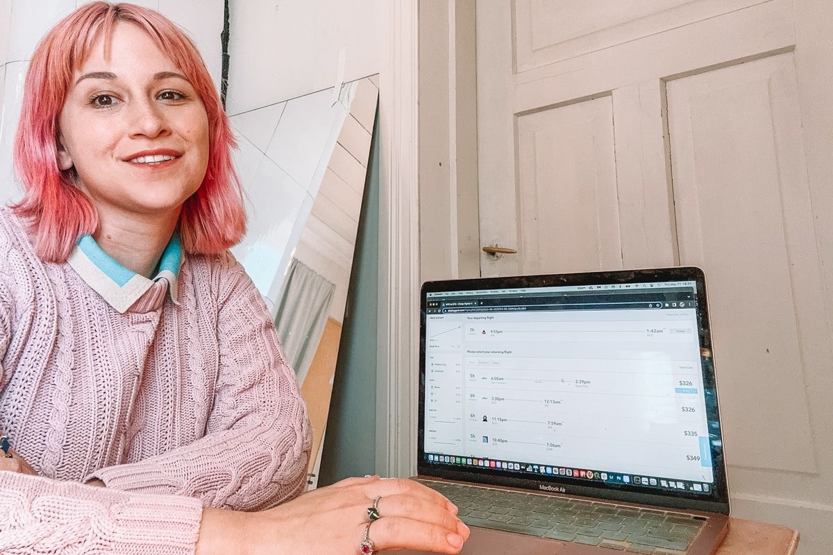A woman with pink hair and a pink sweater sits at a desk and looks at the camera, with the Skiplagged website open on her laptop and a closed white door in the background.