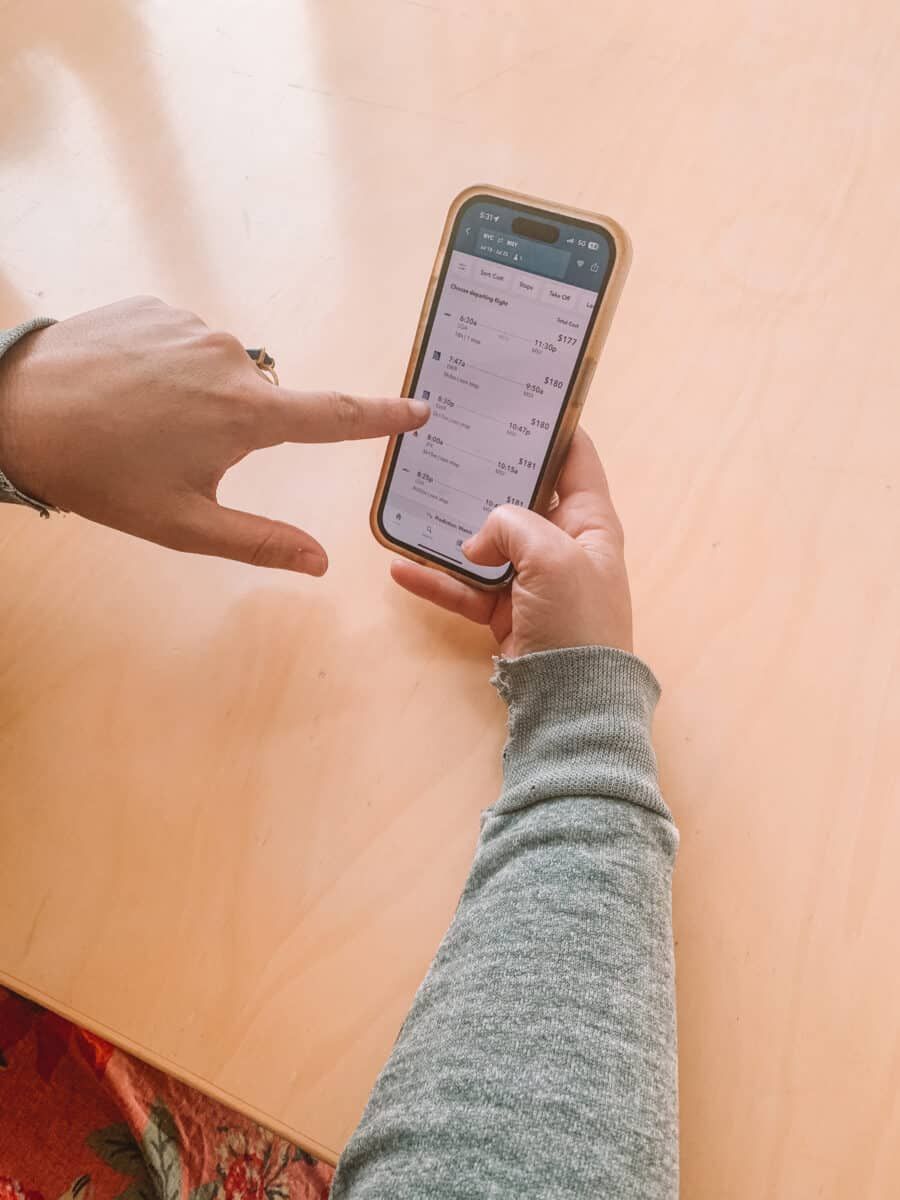 Skiplagged Review: a pair of hands holding an iPhone displaying flight search results on the Skiplagged app and pointing to the screen, resting on a light wood table.
