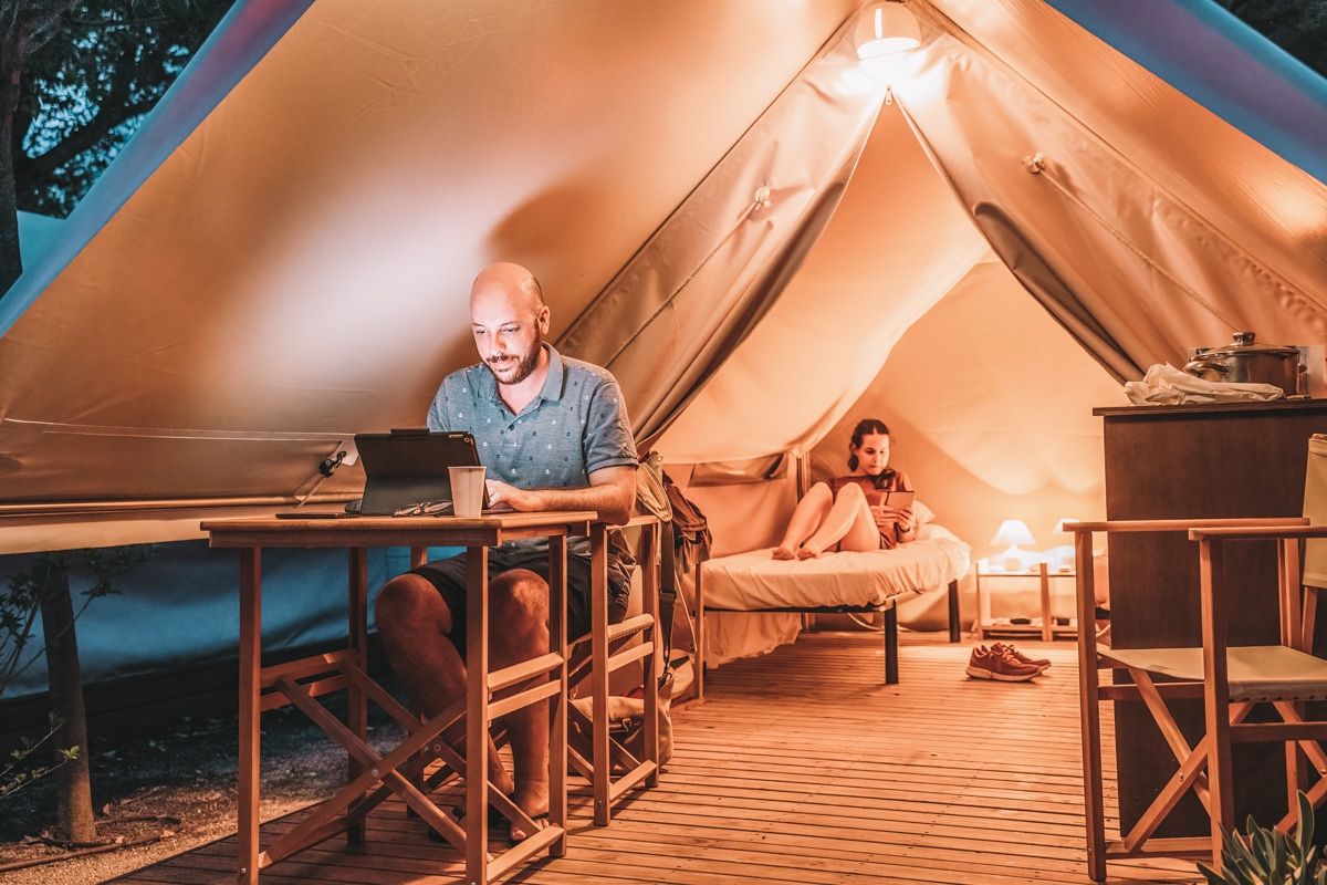 A man sits at a wooden table at dusk at the entrance to a canvas glamping tent working on an iPad, while a woman lounges on a camping cot in the background, read a book.