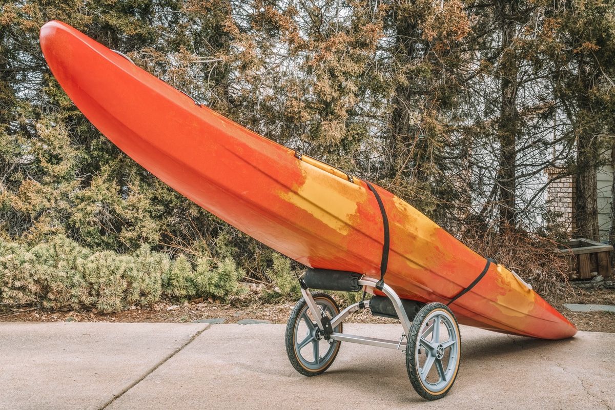 An orange kayak strapped to a small kayak trailer for a bike, sitting on a concrete driveway with pine trees behind it.