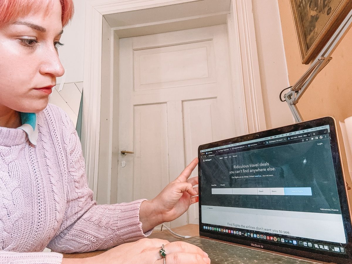 A woman with pink hair and a pink sweater sits at a desk and looks at the Skiplagged homepage on her laptop, with a closed white door in the background.