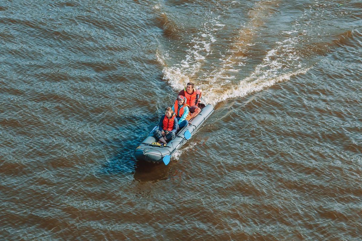 Three people wearing orange life vests seen from above riding in a grey, inflatable, motorized kayak on murky brown water.