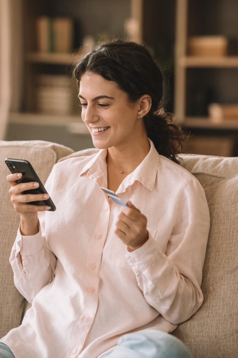 A smiling young woman with curly dark hair sits on a beige couch, browsing one of the best travel deal sites on her phone, while holding up her credit card.