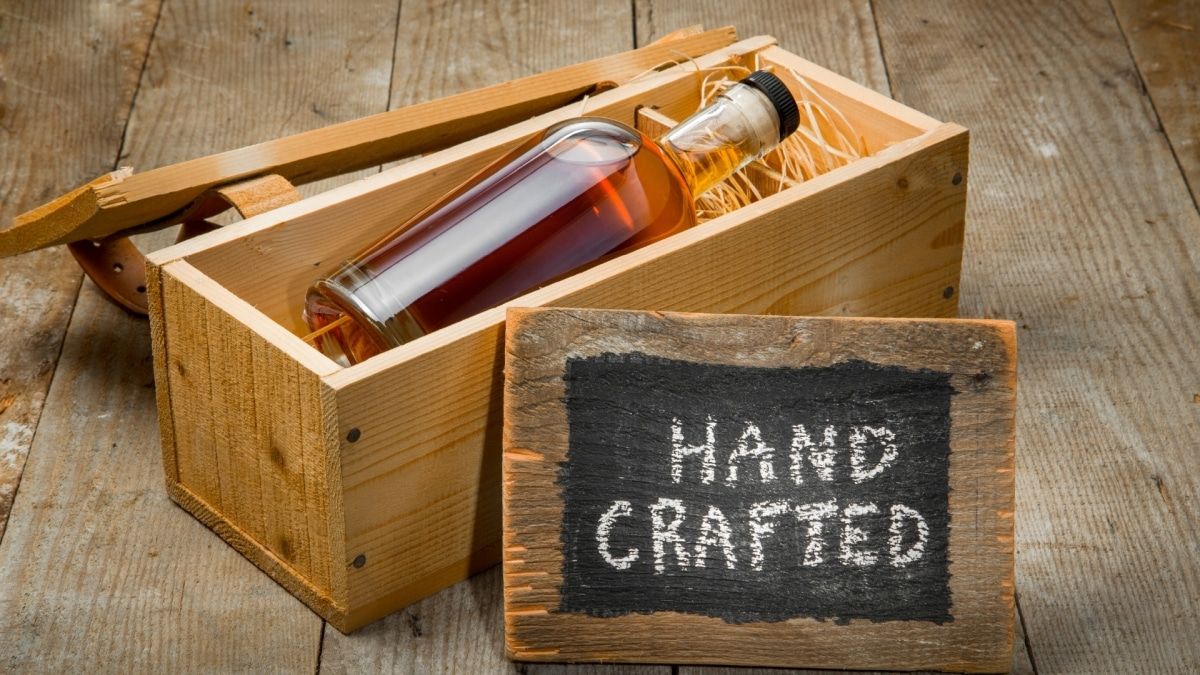 An unlabeled whiskey bottle in a wooden crate gift box, with a rustic blackboard sign that reads, "Hand Crafted."
