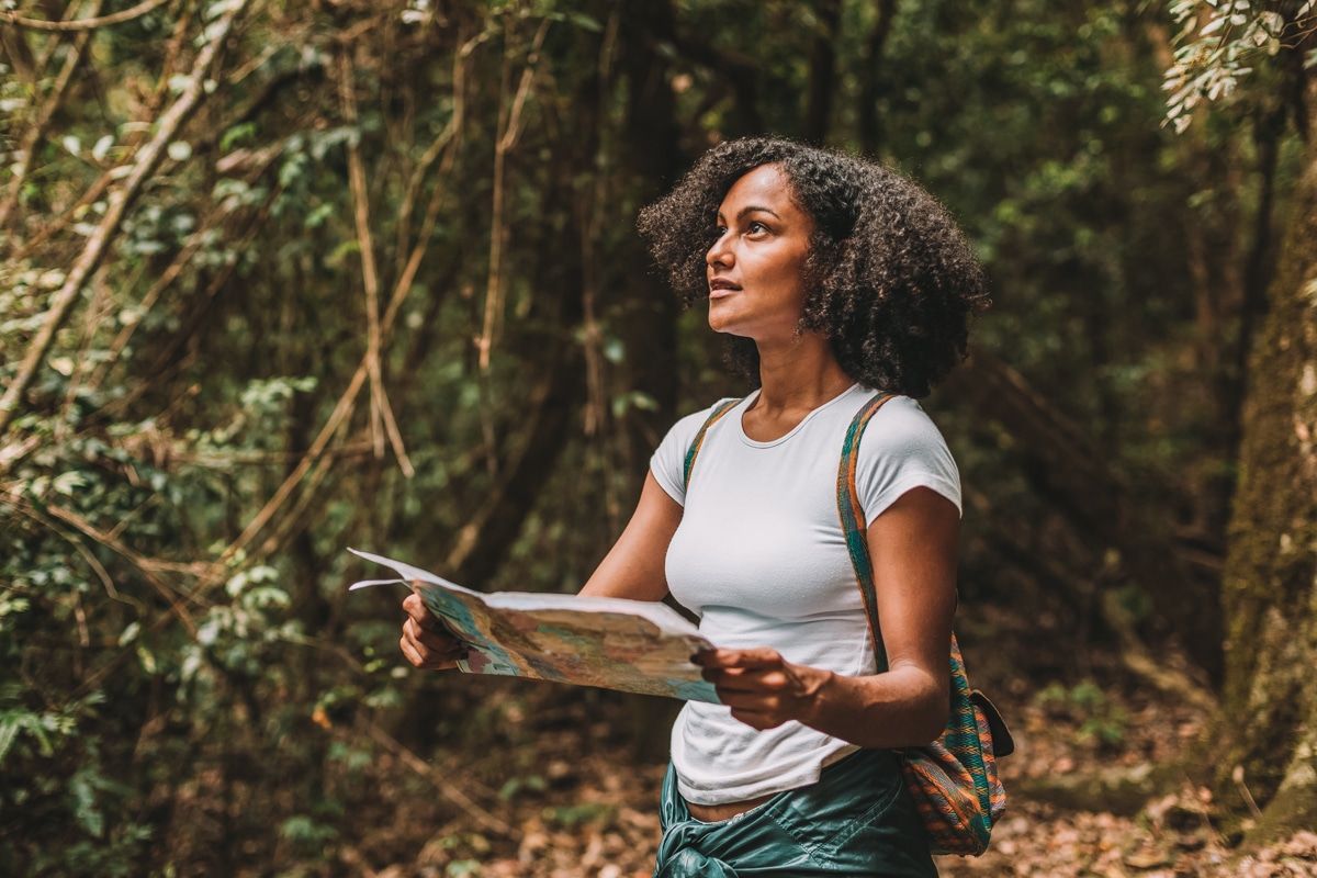 A curly-haired woman in a white t-shirt stands in a dense forest and looks up while holding a map.