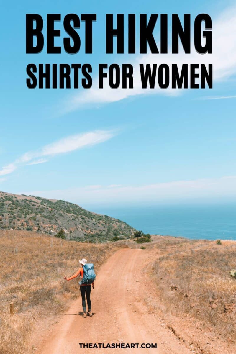 A woman in black leggings and a blue backpack walking down a dry, dusty trail overlooking the ocean with the text overlay, "Best Hiking Shirts for Women."