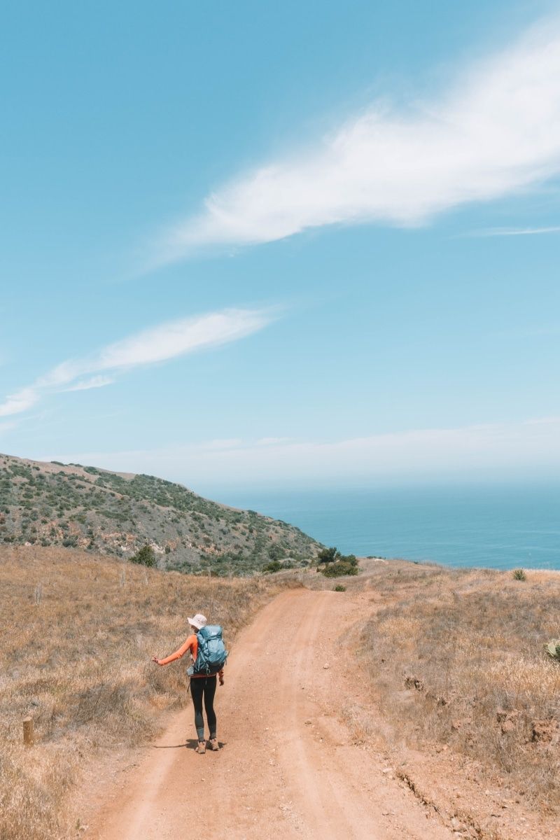 An example of one of the best hiking shirts for women, worn by a woman in black leggings and a blue backpack walking down a dry, dusty trail overlooking the ocean.