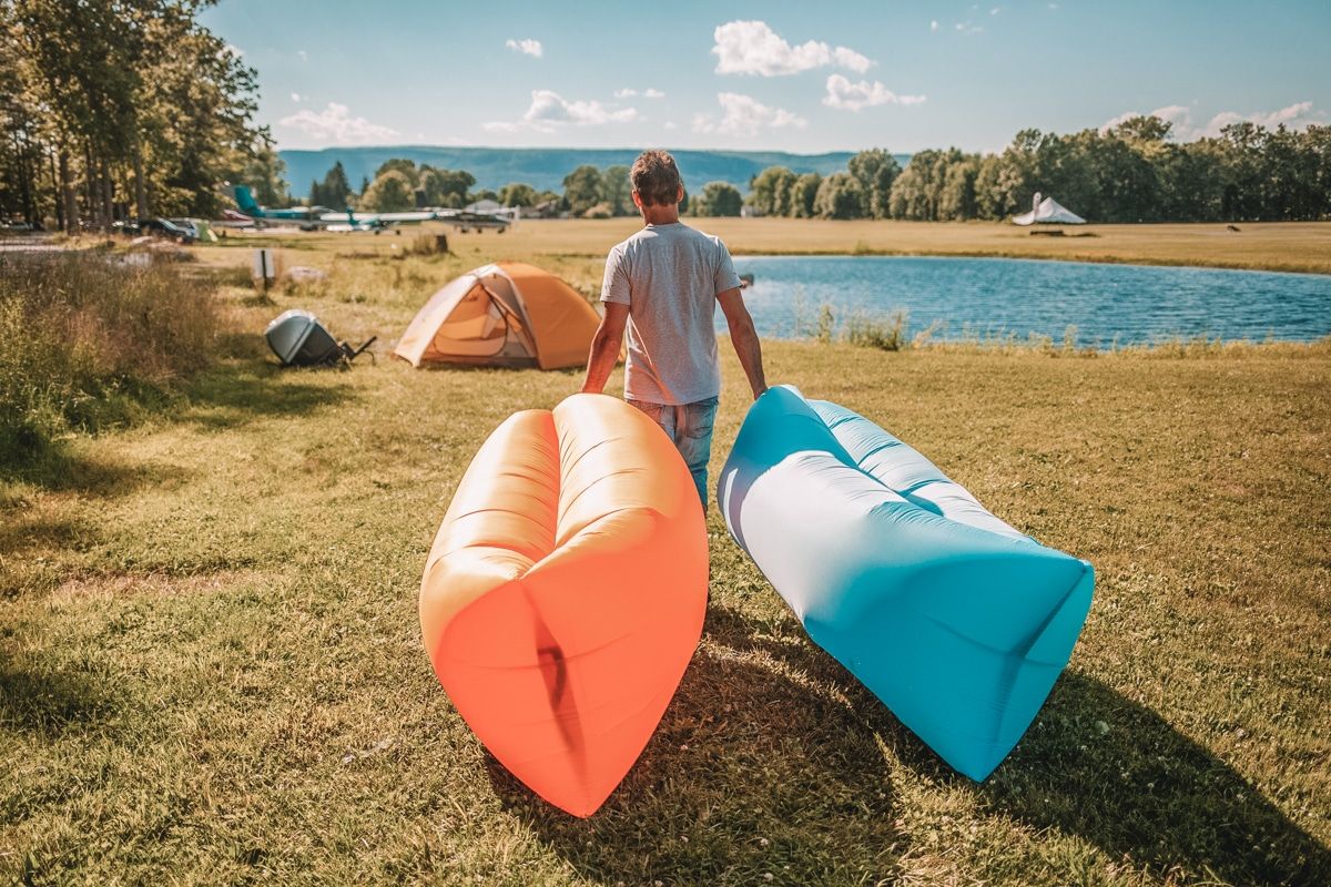 A man drags an orange and a blue inflatable lounger towards a yellow tent pitched next to a small lake.