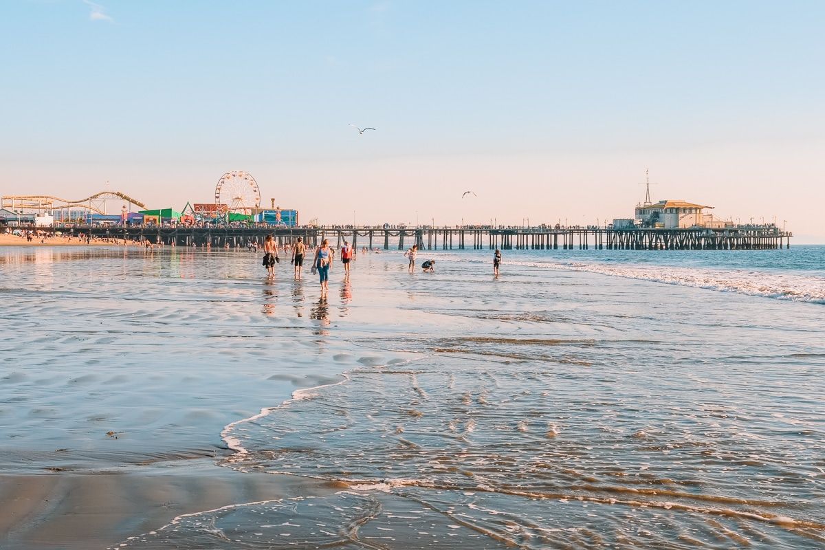 People strolling on the beach at sunset, with the Santa Monica Pier in the background.