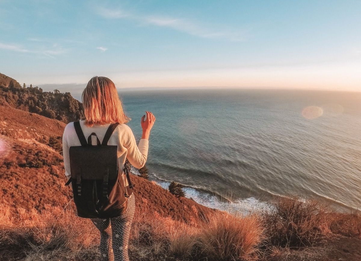 A light-haired woman seen from behind wearing a black backpack stands on a bluff overlooking the ocean at sunset.