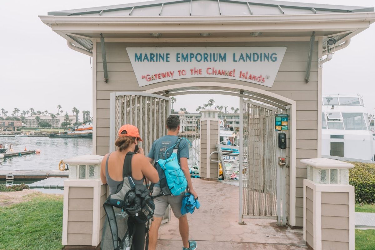 A man and a woman wearing backpacks, seen from behind as they walk through a beige archway with a sign that says, "Marine Emporium Landing," and a harbor visible beyond.