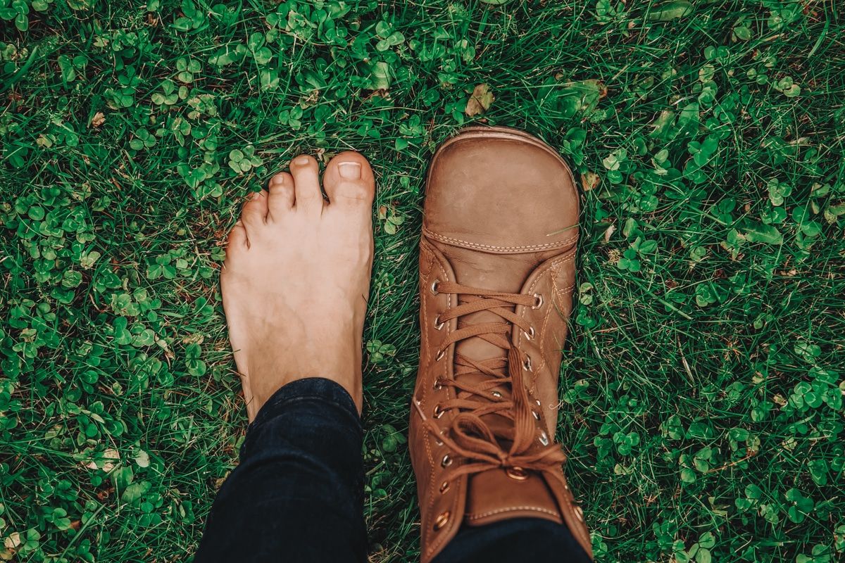 A pair of feet standing on a clover lawn, one bare, and one wearing a brown leather, barefoot-style boot.