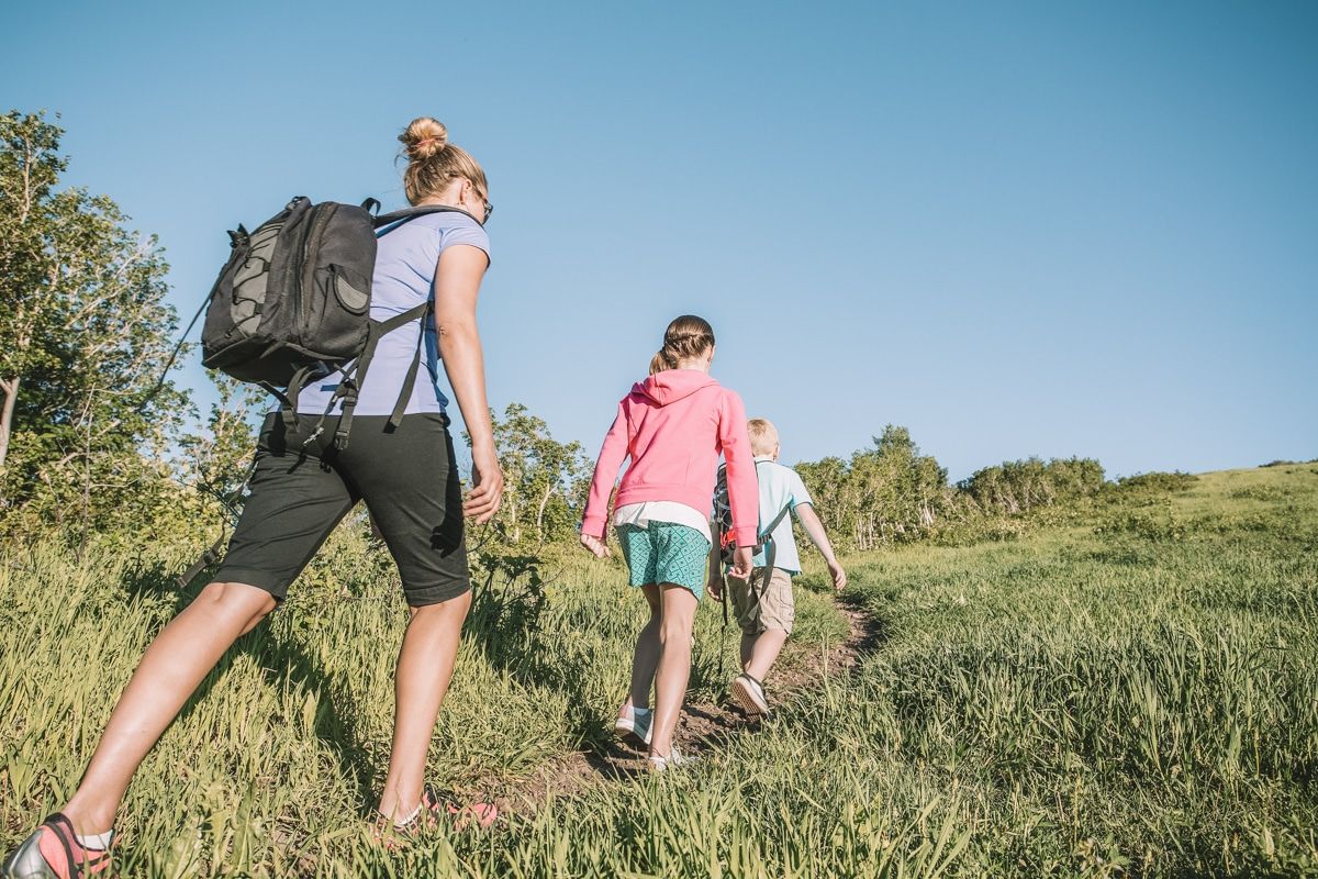 A female hiker wearing a backpack, a purple shirt, and long black shorts hiking behind two children on a grassy trail with a clear blue sky behind them.