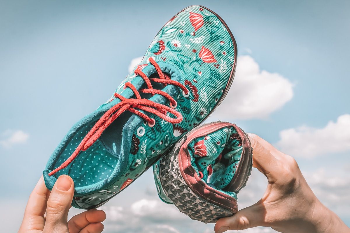 A pair of hands holds up a pair of blue floral print barefoot sneakers, with one rolled up and a cloudy blue sky in the background.