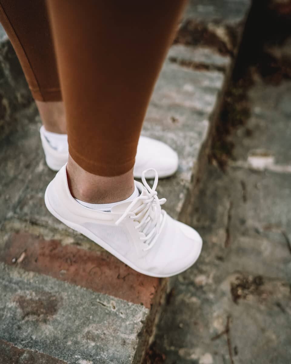 A woman's feet showing off an example of the best casual barefoot shoes in white, standing on brick stairs.